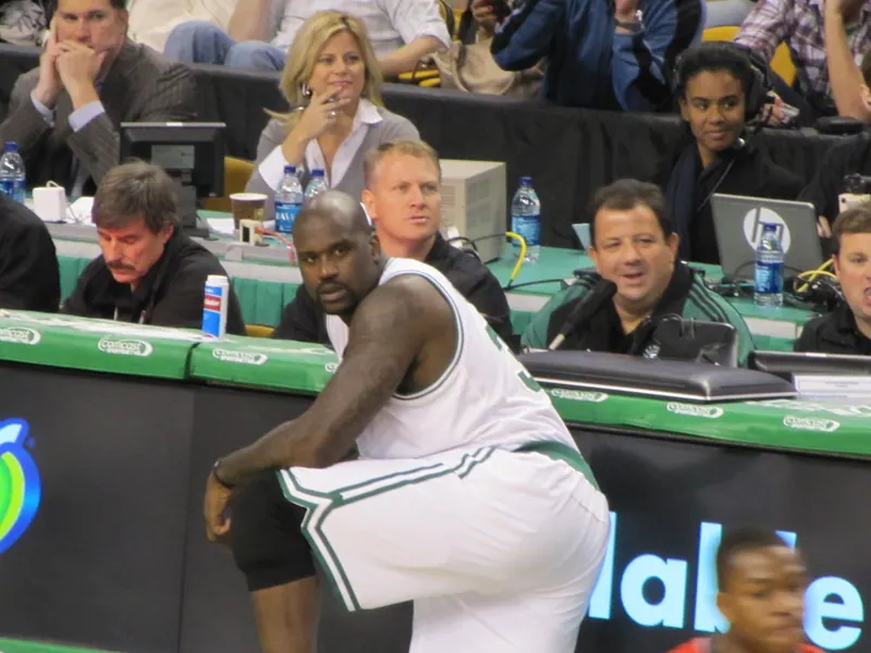 Shaquille O'Neal in a white Boston Celtics uniform crouches at the scorer's table at TD Garden, about to check into one of his first games as a Celtic.
