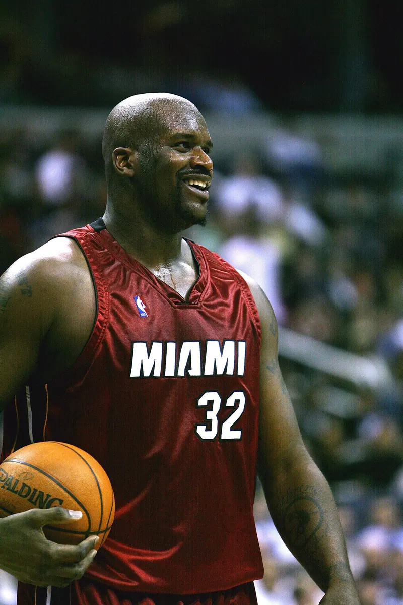 Shaquille O'Neal grinning in a red Miami Heat #32 road jersey, ball on his hip, at a Heat vs. Washington Wizards game.