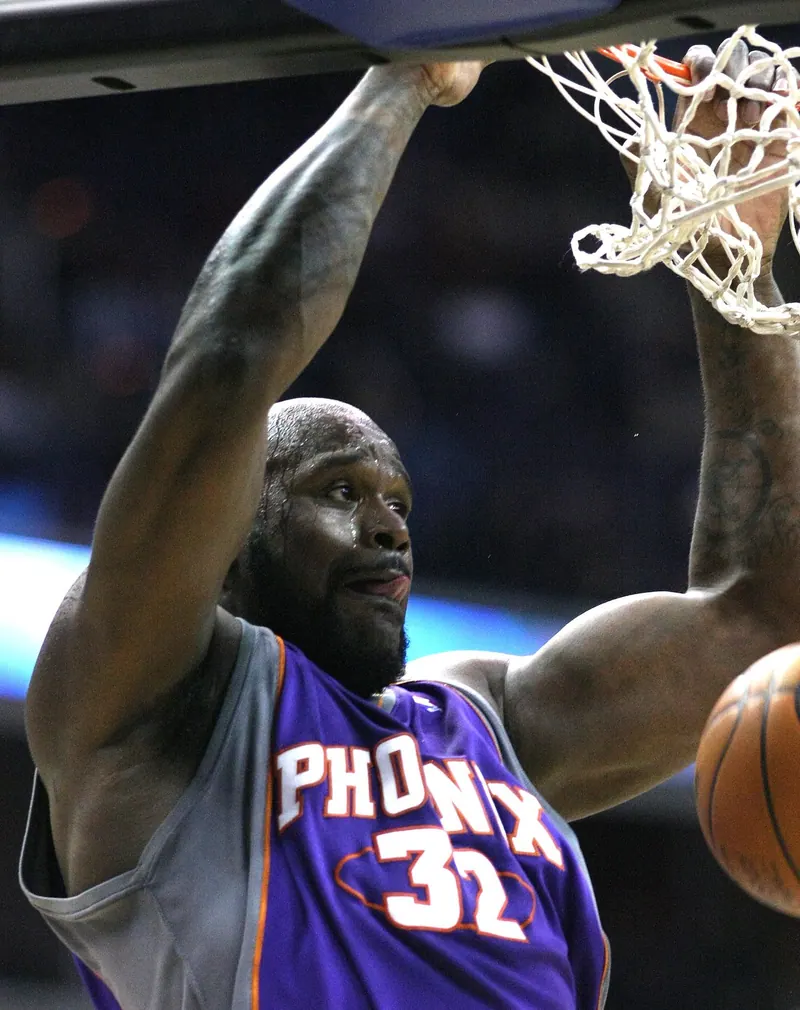 Shaquille O'Neal in a purple Phoenix Suns #32 jersey hangs on the rim after a two-handed dunk, tight action shot.