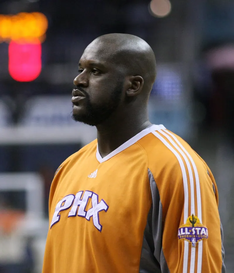 Profile portrait of Shaquille O'Neal in an orange Phoenix Suns 'PHX' warm-up shirt with a 2009 All-Star patch, arena lights behind.